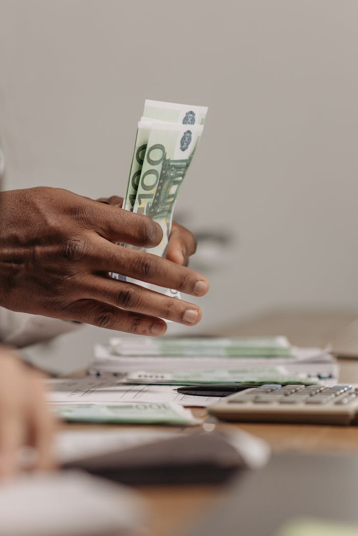 services-04 Close-up of hands counting US dollar bills on a cluttered desk in an office setting.